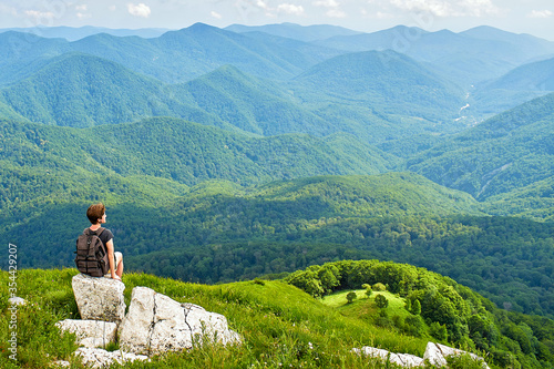 Woman traveler sits on top of a mountain with a beautiful view of the valley. A young female tourist with a backpack on top of a mountain.