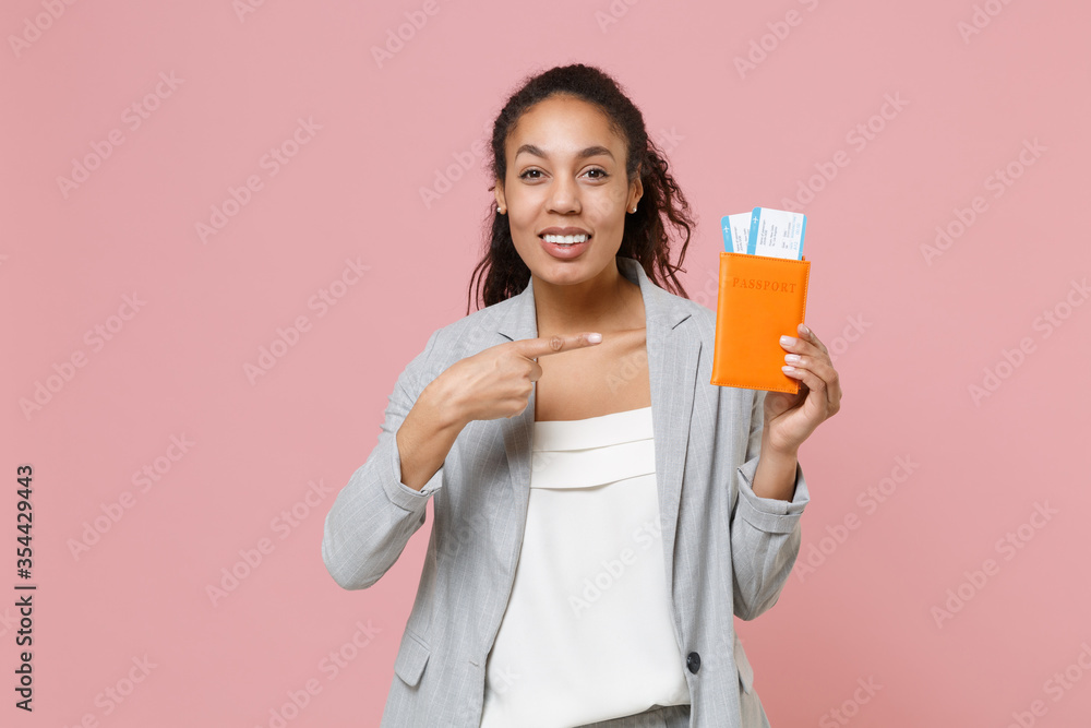 Smiling young african american business woman in grey suit white shirt isolated on pink background. Achievement career wealth business concept. Pointing index finger on passport tickets boarding pass. Smiling young african american business woman in grey suit white shirt isolated on pink background. Achievement career wealth business concept. Pointing index finger on passport tickets boarding pass.
