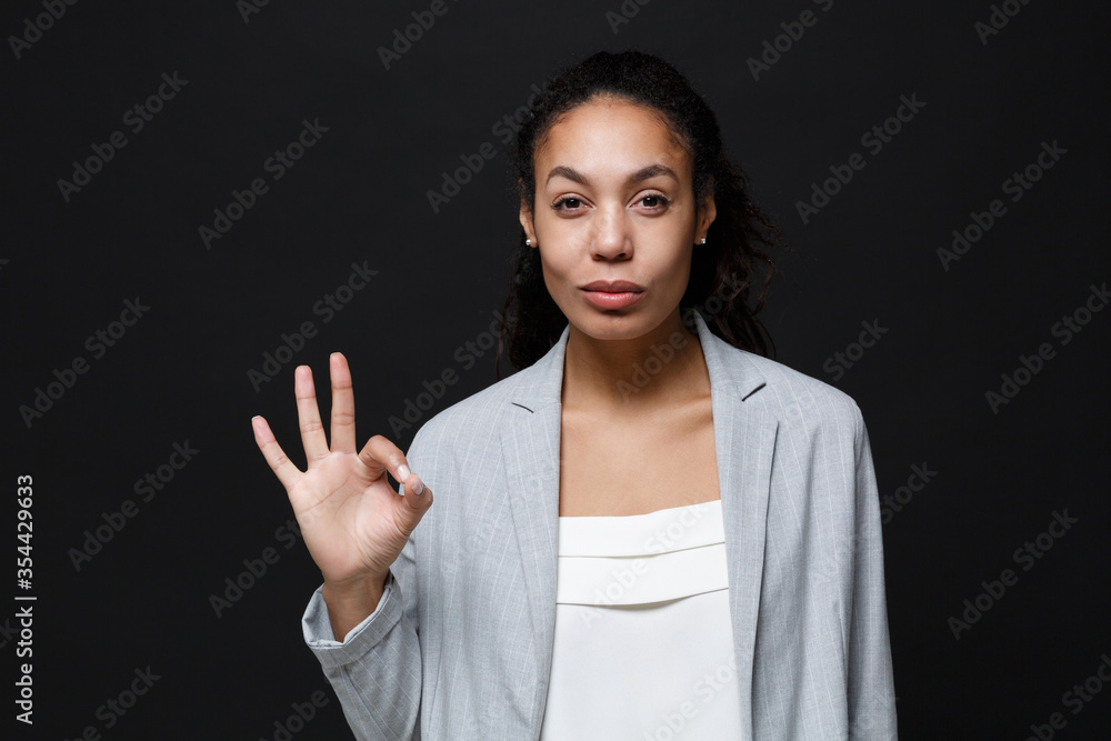 Beautiful young african american business woman in grey suit, white shirt posing isolated on black wall background. Achievement career wealth business concept. Mock up copy space. Showing OK gesture. Beautiful young african american business woman in grey suit, white shirt posing isolated on black wall background. Achievement career wealth business concept. Mock up copy space. Showing OK gesture.