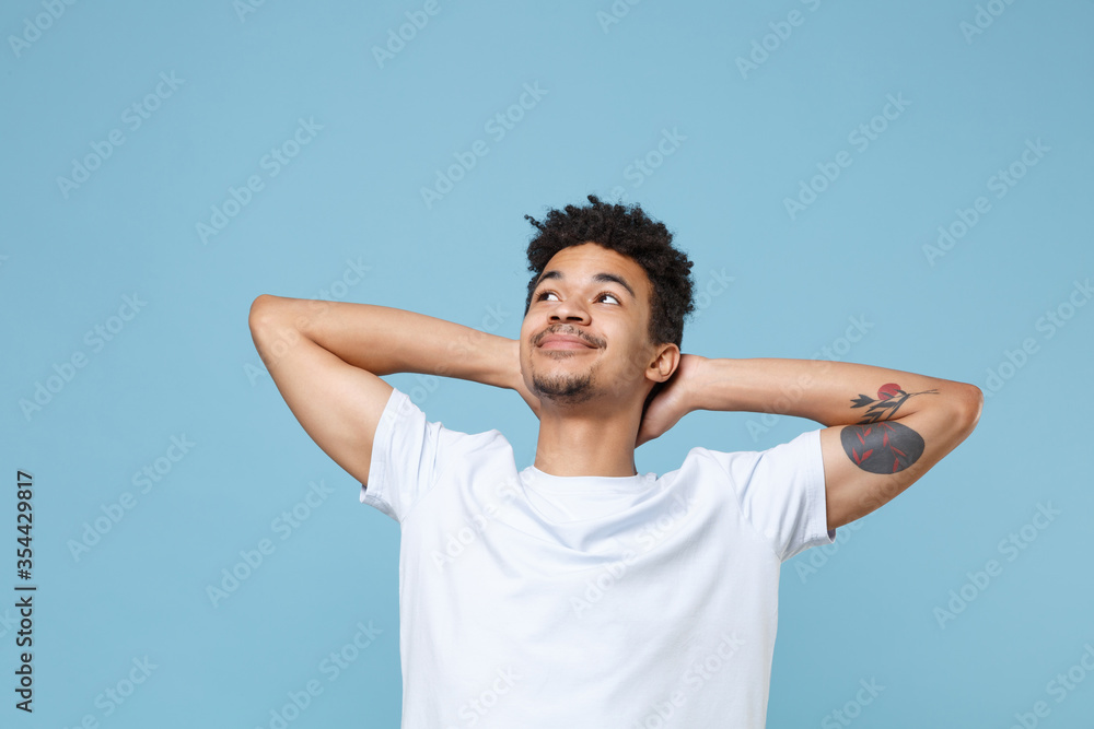 Smiling young african american guy in casual white t-shirt posing isolated on pastel blue background studio portrait. People lifestyle concept. Mock up copy space. Looking up with hands behind head. Smiling young african american guy in casual white t-shirt posing isolated on pastel blue background studio portrait. People lifestyle concept. Mock up copy space. Looking up with hands behind head.