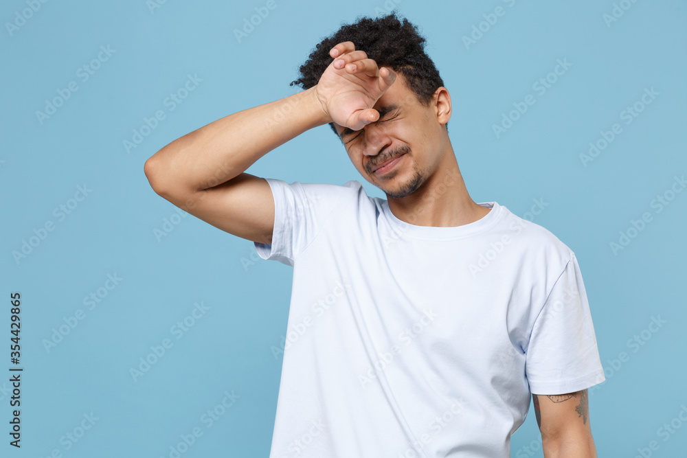 Fototapeta premium Exhausted tired young african american guy in casual white t-shirt posing isolated on pastel blue background studio. People lifestyle concept. Mock up copy space. Put hand on head keeping eyes closed.