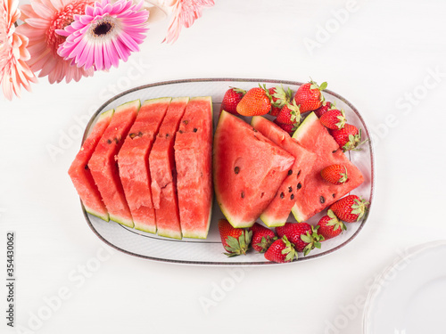 Watermelon slices with strawberries on ceramic tray on white wooden table with pink flowers. Fresh summer snack
