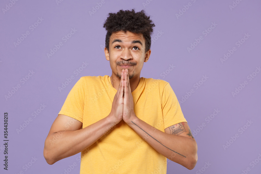Pensive young african american guy in casual yellow t-shirt posing isolated on pastel violet background studio portrait. People lifestyle concept. Mock up copy space. Holding hands folded in prayer. Pensive young african american guy in casual yellow t-shirt posing isolated on pastel violet background studio portrait. People lifestyle concept. Mock up copy space. Holding hands folded in prayer.
