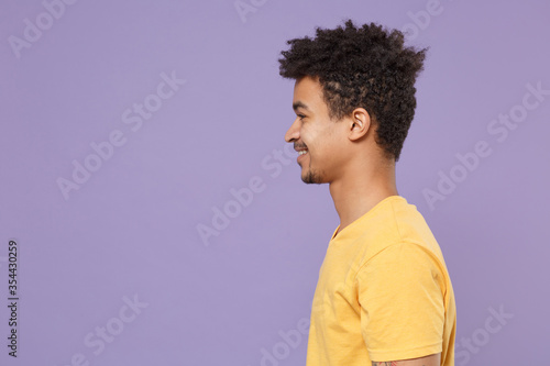 Side view of smiling young african american guy in casual yellow t-shirt posing isolated on pastel violet wall background studio portrait. People lifestyle concept. Mock up copy space. Looking aside.
