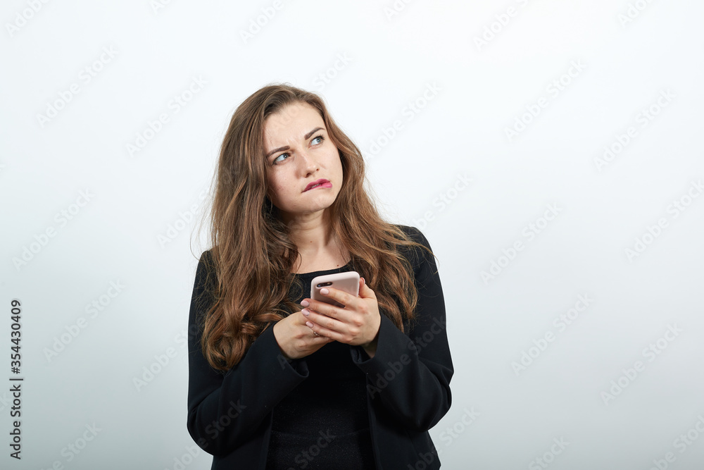 Young Attractive Woman Brunette In In Black T-Shirt And Sweater On White Background, Focused Female Holding A Phone In Her Hand. Communication Via Smartphone, The Communication People Young Attractive Woman Brunette In In Black T-Shirt And Sweater On White Background, Focused Female Holding A Phone In Her Hand. Communication Via Smartphone, The Communication People