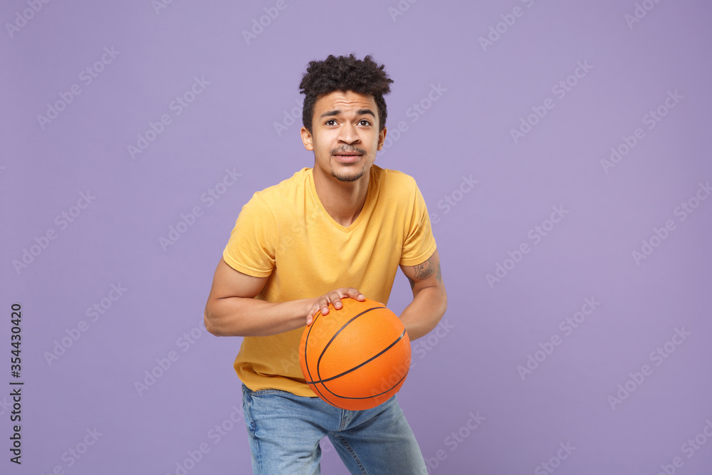 Young african american guy basketball player in casual yellow t-shirt posing isolated on pastel violet background studio. People emotions, sport leisure lifestyle concept. Play basketball with ball. Young african american guy basketball player in casual yellow t-shirt posing isolated on pastel violet background studio. People emotions, sport leisure lifestyle concept. Play basketball with ball.