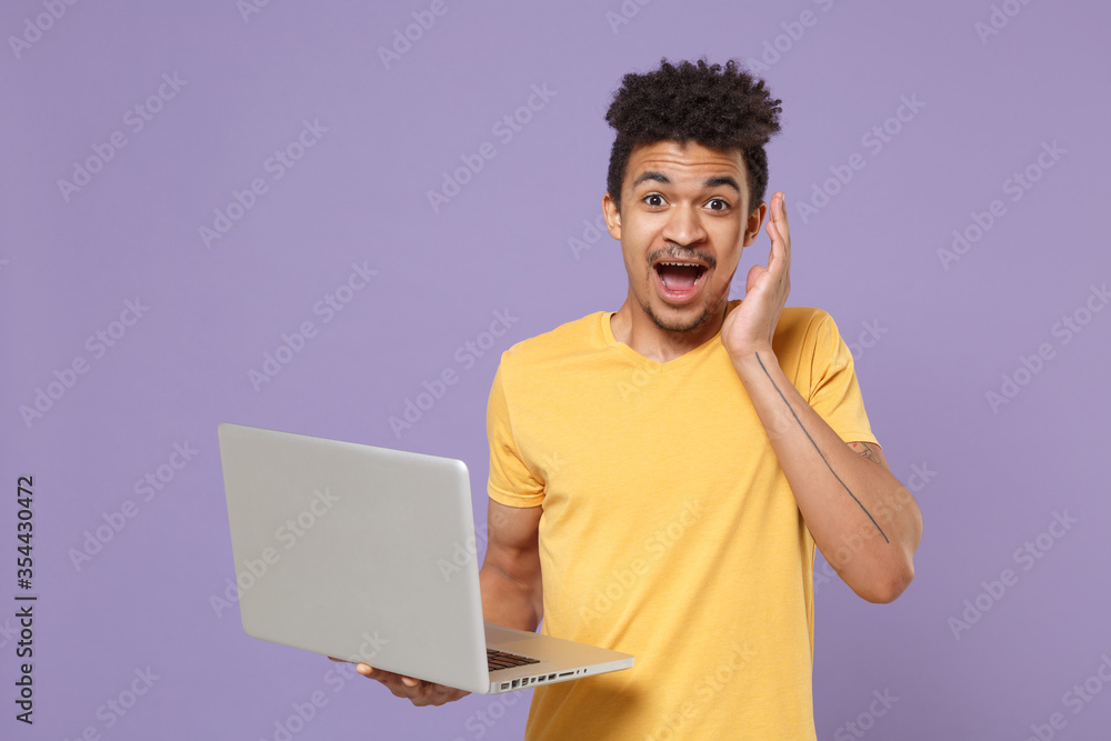 Shocked young african american guy in casual yellow t-shirt posing isolated on pastel violet background. People lifestyle concept. Mock up copy space. Working on laptop pc computer, put hand on cheek. Shocked young african american guy in casual yellow t-shirt posing isolated on pastel violet background. People lifestyle concept. Mock up copy space. Working on laptop pc computer, put hand on cheek.