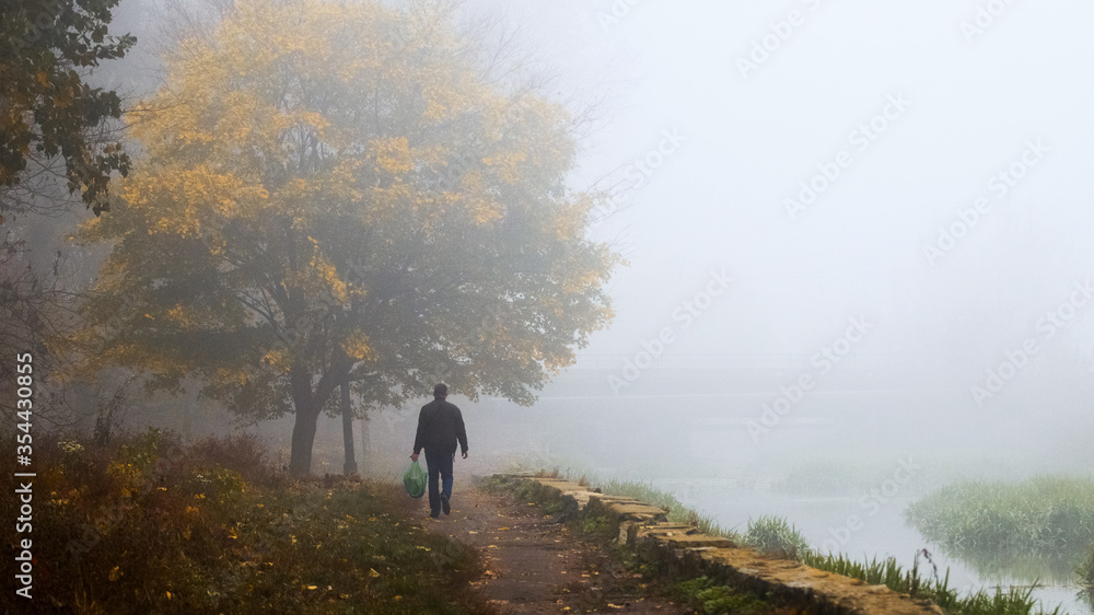 A man walks in the autumn park by the lake on a foggy morning