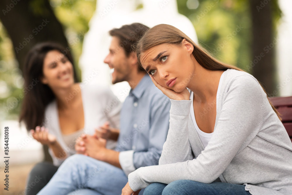 Depressed Girl Sitting Next To Happy Friends In Park Outside
