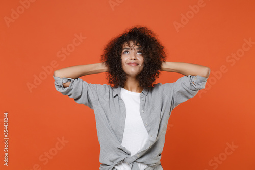 Smiling young african american woman girl in gray casual clothes isolated on orange wall background studio portrait. People lifestyle concept. Mock up copy space. Looking up with hands behind head.