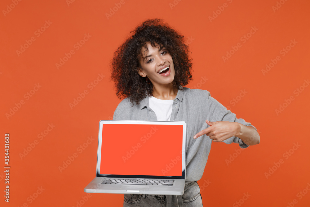 Naklejka premium Funny young african american woman in gray casual clothes isolated on orange background. People lifestyle concept. Mock up copy space. Point index finger on laptop pc computer with blank empty screen.