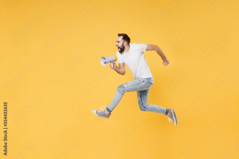 Crazy young bearded man guy in white casual t-shirt posing isolated on yellow wall background studio portrait. People lifestyle concept. Mock up copy space. Jumping like running, scream in megaphone.