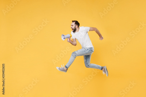 Crazy young bearded man guy in white casual t-shirt posing isolated on yellow wall background studio portrait. People lifestyle concept. Mock up copy space. Jumping like running  scream in megaphone.