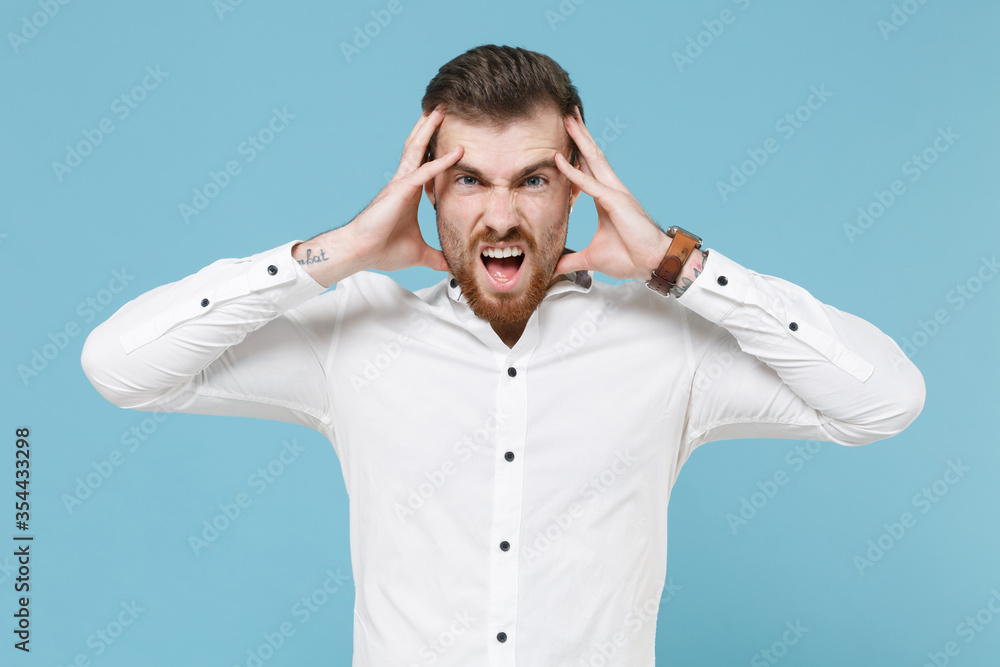 Naklejka premium Displeased tired young bearded man guy 20s in white classic shirt isolated on pastel blue background studio portrait. People sincere emotions lifestyle concept. Mock up copy space. Put hands on head.