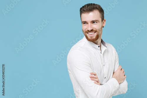 Smiling young bearded man guy 20s in white classic shirt isolated on pastel blue wall background studio portrait. People lifestyle concept. Mock up copy space. Holding hands crossed, looking aside.