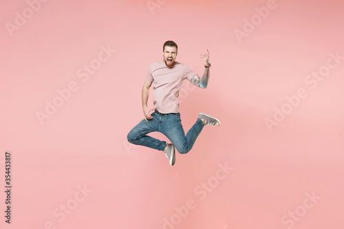 Crazy young bearded tattooed man guy in pastel casual t-shirt posing isolated on pink wall background studio. People lifestyle concept. Mock up copy space. Jumping, holding hands like playing guitar.