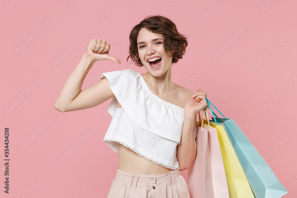 Laughing young woman girl in summer clothes hold package bag with purchases isolated on pink background studio portrait. Shopping discount sale concept. Mock up copy space. Pointing thumb on himself.