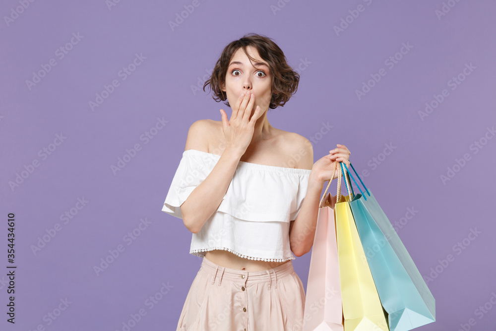Shocked young woman girl in summer clothes hold package bag with purchases isolated on pastel violet background in studio. Shopping discount sale concept. Mock up copy space. Covering mouth with hand. Shocked young woman girl in summer clothes hold package bag with purchases isolated on pastel violet background in studio. Shopping discount sale concept. Mock up copy space. Covering mouth with hand.