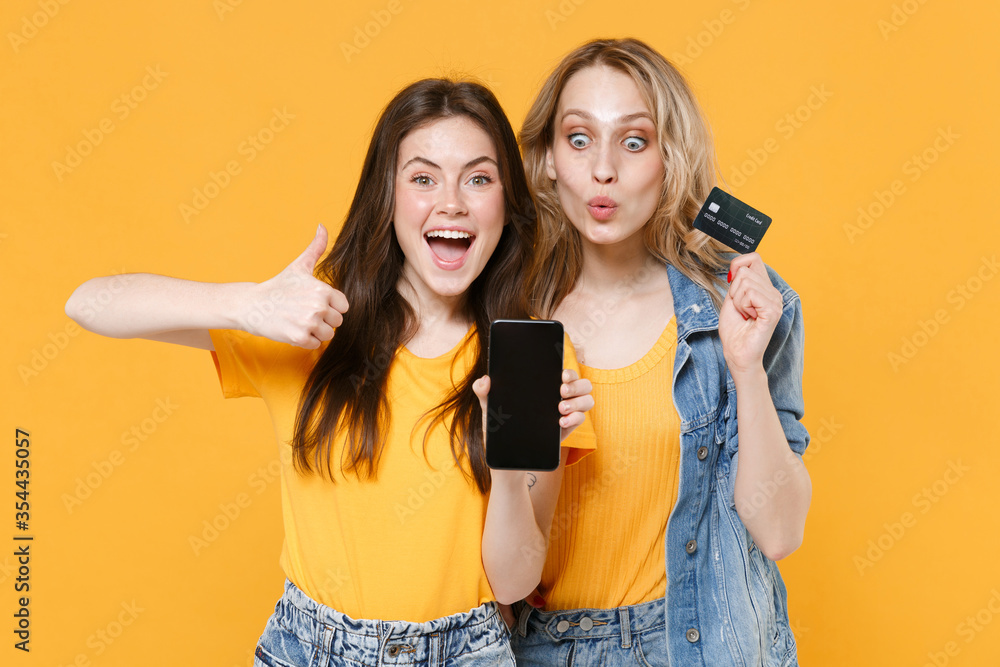 Two excited women girls friends in casual t-shirts denim clothes isolated on yellow background. People lifestyle concept. Hold mobile phone with blank empty screen credit bank card, showing thumb up. Two excited women girls friends in casual t-shirts denim clothes isolated on yellow background. People lifestyle concept. Hold mobile phone with blank empty screen credit bank card, showing thumb up.