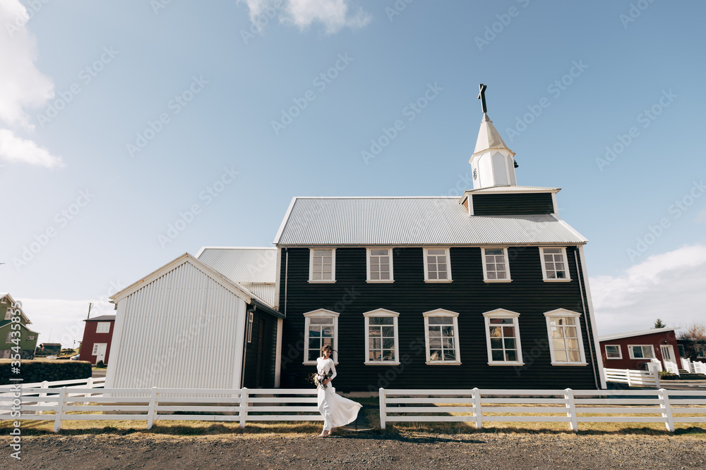 The bride in a white dress with a bouquet in her hands, goes against the background of wooden windows of a black church and a white fence. Destination Iceland wedding. 