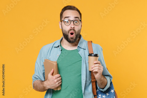 Shocked young man student in glasses backpack hold books isolated on yellow background studio. Education in high school university college concept. Mock up copy space. Hold paper cup of coffee or tea.