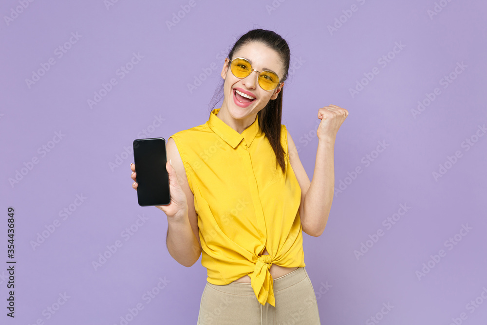 Joyful young brunette woman in yellow casual shirt posing isolated on violet background. People lifestyle concept. Mock up copy space. Hold mobile phone with blank empty screen doing winner gesture. Joyful young brunette woman in yellow casual shirt posing isolated on violet background. People lifestyle concept. Mock up copy space. Hold mobile phone with blank empty screen doing winner gesture.