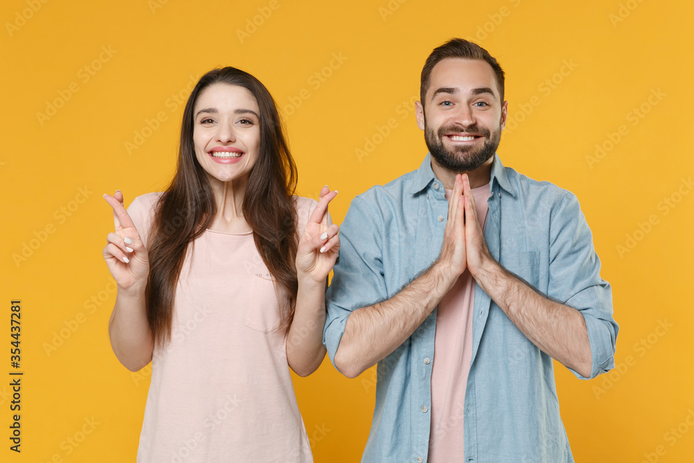 Young couple friends guy girl in casual clothes isolated on yellow background. People lifestyle concept. Waiting for special moment, keeping fingers crossed making wish holding hands folded in prayer.