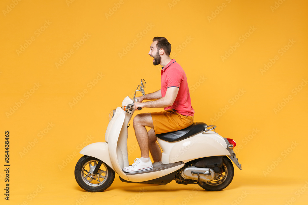 Side view of shocked young bearded man guy in casual summer clothes driving moped isolated on yellow wall background studio portrait. Driving motorbike transportation concept. Mock up copy space. Side view of shocked young bearded man guy in casual summer clothes driving moped isolated on yellow wall background studio portrait. Driving motorbike transportation concept. Mock up copy space.