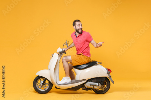 Side view of excited young bearded man guy in casual summer clothes driving moped isolated on yellow background. Driving motorbike transportation concept. Mock up copy space. Point index finger aside. © ViDi Studio