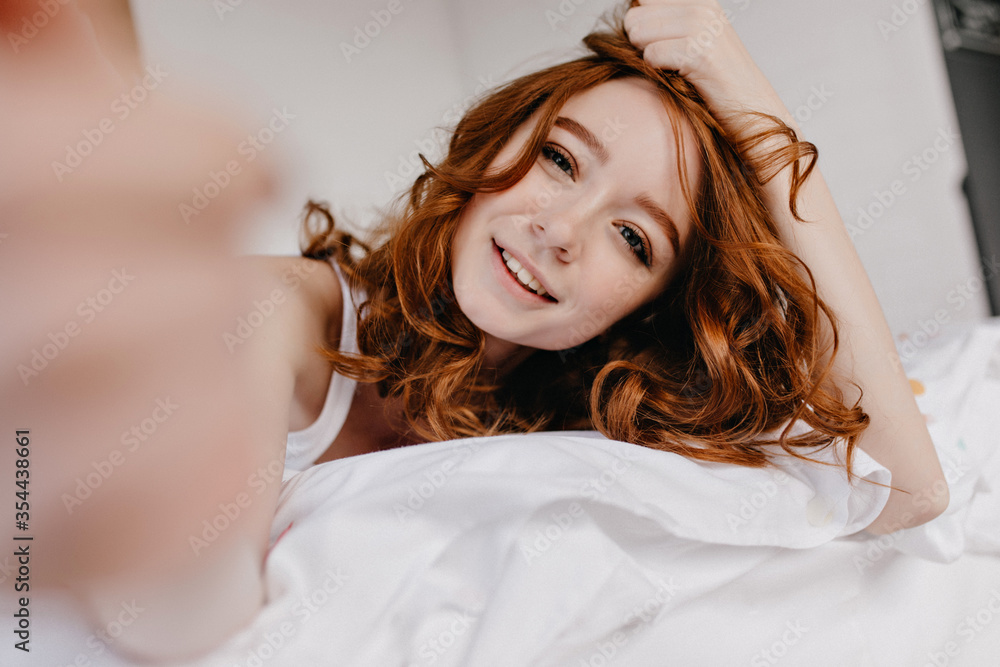 Fototapeta premium Spectacular white girl with ginger hair relaxing in morning. Indoor photo of lovely caucasian young lady making selfie in bed.