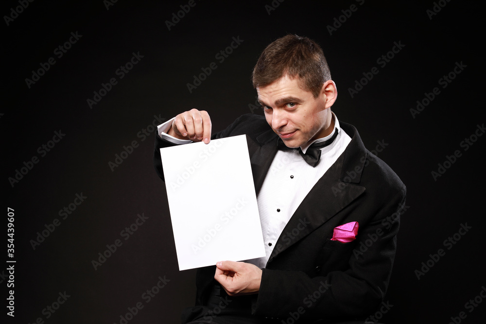Young handsome man in black tuxedo holding clear white paper with copy space isolated on black background Young handsome man in black tuxedo holding clear white paper with copy space isolated on black background