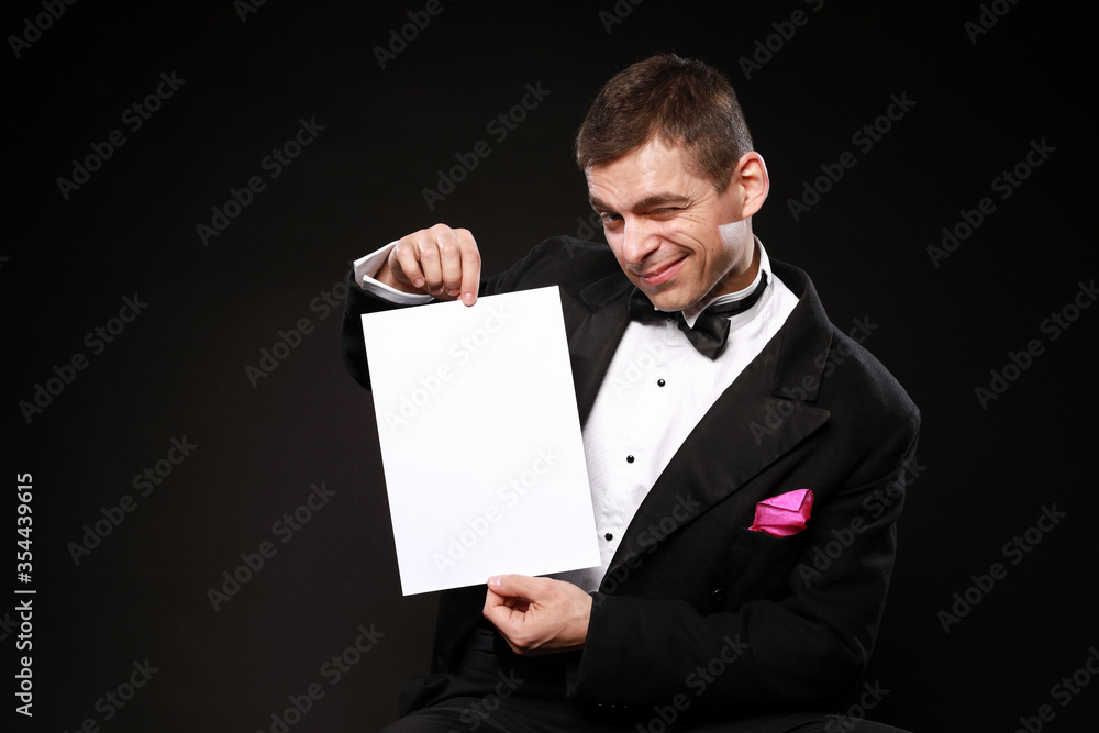 Young handsome man in black tuxedo holding clear white paper with copy space isolated on black background Young handsome man in black tuxedo holding clear white paper with copy space isolated on black background
