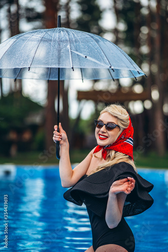 Pin up styled blonde in sunglasses holding umbrella and have fun in the swimming pool