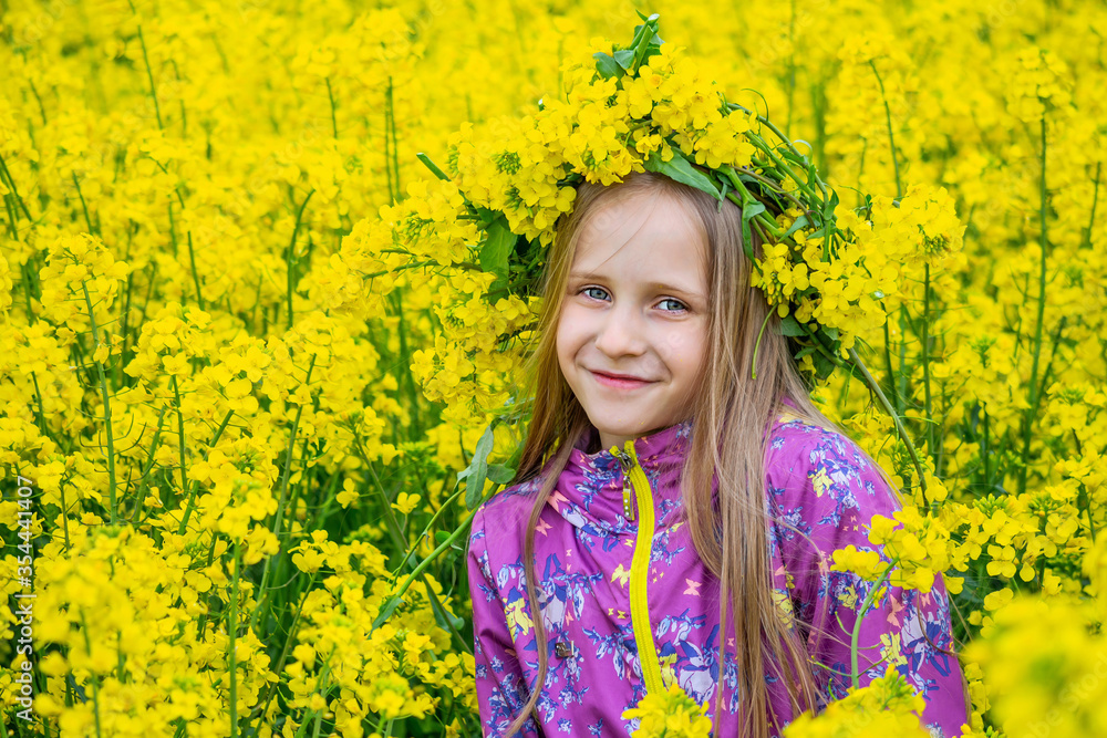 Happy girl with long light hair and grey eyes is standing in the field of yellow flowers. The wreath from canola is beautify the head. Rural landscape of blooming rapeseed. Happy girl with long light hair and grey eyes is standing in the field of yellow flowers. The wreath from canola is beautify the head. Rural landscape of blooming rapeseed.