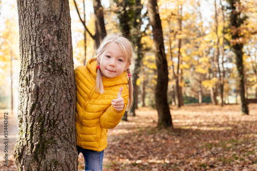 Portrait of child positive 5-6 year old girl in  yellow jacket looks out from behind  tree and shows thumbs up on walk in the park on sunny autumn day  Kids outdoors, hiking with his family in fall © tgordievskaya