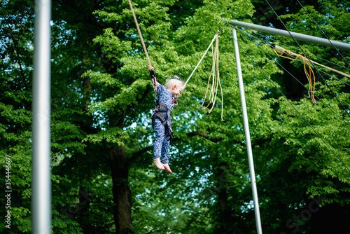 The girl is jumping on a bungee trampoline. A child with insurance and stretchable rubber bands hangs against the sky. The concept of happy childhood and games in the amusement park