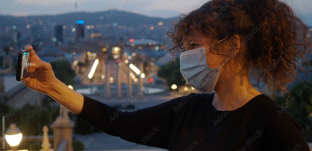 Fototapeta premium Tourist woman with mask on her face takes a selfie at night in the city. New normal