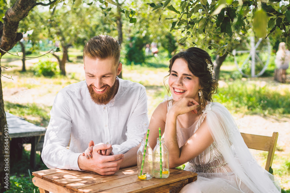 Attractive couple newlyweds, happy and joyful moment. bride and groom sit at table set for two in woods. Concept romantic date. Wedding couple sitting in cafe table and lovingly look at each other Attractive couple newlyweds, happy and joyful moment. bride and groom sit at table set for two in woods. Concept romantic date. Wedding couple sitting in cafe table and lovingly look at each other