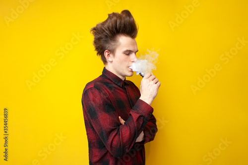hipster guy smokes vape on yellow isolated background  man exhales a cloud of smoke and holds an electronic cigarette