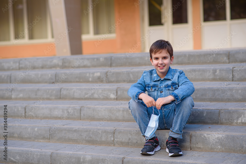 Smiling schoolboy sits on stairs outdoors in schoolyard with protective mask in hands. End of pandemic coronavirus 2020.