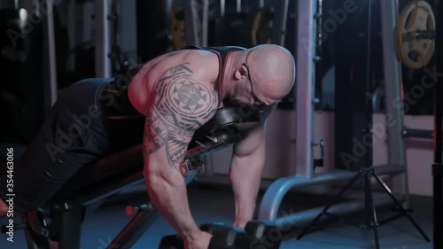 Professional bodybuilder trains back deltoid muscle on a bench with dumbbells in a sports club.