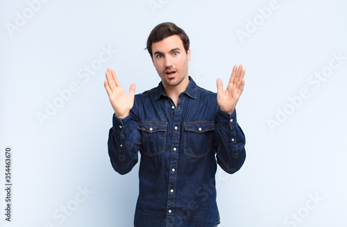 young handsome man looking shocked and astonished, with jaw dropped in surprise when realizing something unbelievable against blue wall photo