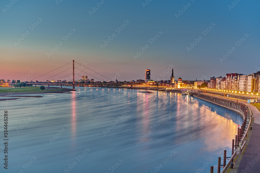 Evening photo of the Rhine and the illuminated promenade of the city of Düsseldorf. In the background a bridge spans the Rhine. One bank side is greened.