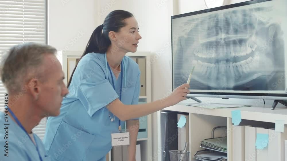Medium shot of young Caucasian female prosthodontist discussing x-ray image on big screen with her male colleague Medium shot of young Caucasian female prosthodontist discussing x-ray image on big screen with her male colleague