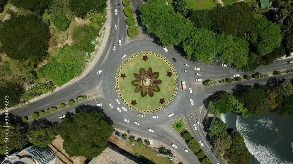 Aerial lockdown shot of vehicles at roundabout amidst trees on sunny day, drone flying over cars in city - Yangon, Myanmar