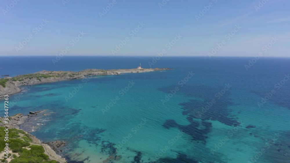 Favaritx Lighthouse Seen On The Distance. A Crystal Clear Emerald Water Of The Ocean Surrounded By Lush Meadow In Menorca, Balearic Islands, Spain. - aerial drone backward Favaritx Lighthouse Seen On The Distance. A Crystal Clear Emerald Water Of The Ocean Surrounded By Lush Meadow In Menorca, Balearic Islands, Spain. - aerial drone backward