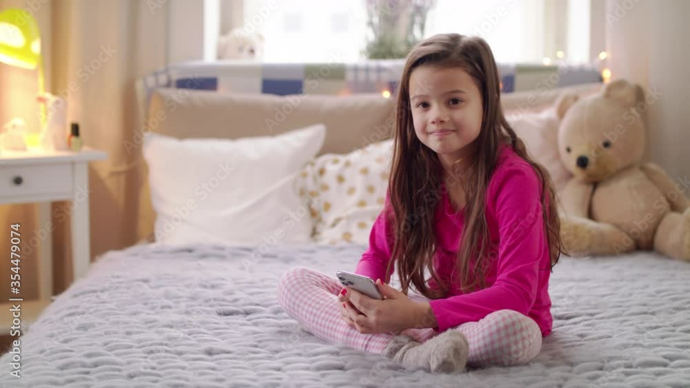 Portrait of small girl with smartphone sitting on bed, danger of addiction concept. Portrait of small girl with smartphone sitting on bed, danger of addiction concept.