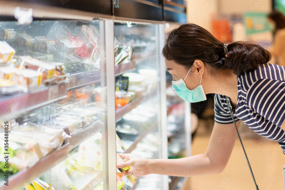 Asian young adult woman walking in the grocery store and looking a goods on shelf during the Coronavirus or COVID-19 outbreak. Woman wearing a protective mask looking for food in supermarket close up Asian young adult woman walking in the grocery store and looking a goods on shelf during the Coronavirus or COVID-19 outbreak. Woman wearing a protective mask looking for food in supermarket close up
