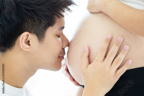 Asian lovely husband and wife portrait, his wife is in pregnancy. Husband tenderly hug and kiss his wife on the bed with care. Husband embracing his pregnant wife and making their hands in heart shape