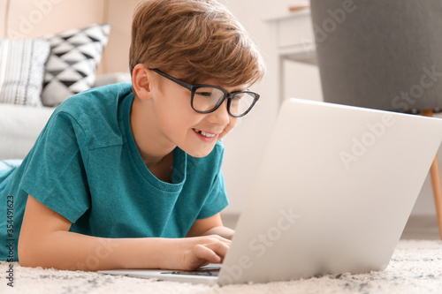 Smiling young boy wearing glasses lying on floor using laptop computer for on...